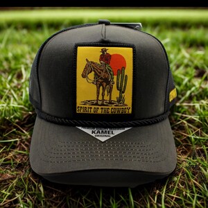 May include: A black trucker hat with a flat peak and a mesh back. The front has a yellow patch with a cowboy on a horse, a red sun, and the text "Spirit of the Cowboy."