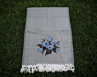 May include: A gray rectangular textile with a floral design. The design features blue flowers with black and green leaves. The bottom edge has a white fringe. The textile is displayed on a green grassy surface.