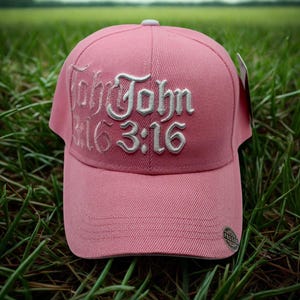 May include: A pink baseball cap with the text "John 3:16" embroidered in white script. The cap has a curved brim and a small, circular emblem on the side. The cap is set against a background of green grass.