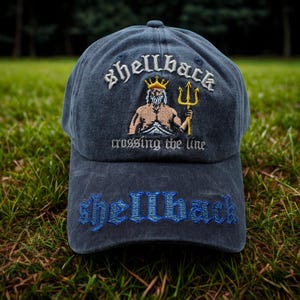 Może przedstawiać: Granatowa czapka bejsbolówka z napisem "shellback" wyszytym białą czcionką nad ilustracją muskularnej postaci z trójzębem. Fraza "crossing the line" znajduje się pod ilustracją, a napis "shellback" jest wyszyty na niebiesko na daszku.
