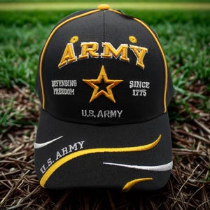 May include: Black baseball cap with gold and white embroidered details. The cap features the word "ARMY" in gold, a gold star, and the text "DEFENDING FREEDOM SINCE 1775 U.S. ARMY". The brim has curved gold and white accents.