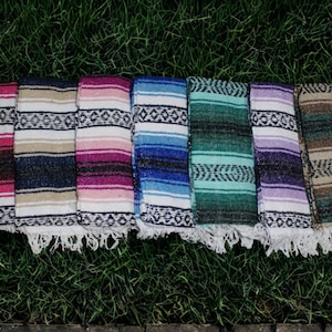 May include: A collection of seven rectangular woven blankets featuring horizontal stripes in various colors including pink, tan, blue, teal, purple, and brown, each accented with black geometric patterns and finished with white fringe at the ends.