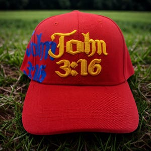 May include: A red baseball cap with the text "John 3:16" embroidered in yellow and blue. The hat is sitting on green grass.