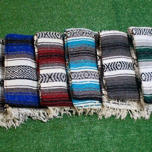 May include: A collection of eight folded rectangular blankets featuring woven patterns with stripes and geometric diamond motifs. Each blanket displays a unique color combination including red, gray, blue, white, turquoise, black, and green, all finished with fringed edges.
