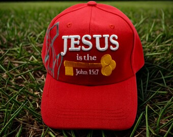 May include: A red baseball cap with the text "JESUS is the" and "John 15:7" embroidered in white and gold. The cap features a key design and a gray decorative element on the side. The cap is sitting on green grass.
