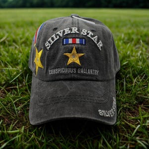 May include: A charcoal gray baseball cap with the words "SILVER STAR" embroidered in white. The cap features a gold star and a ribbon emblem. The text "CONSPICUOUS GALLANTRY" is also embroidered on the cap.