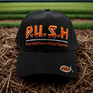 May include: Black baseball cap with the embroidered text "P.U.S.H. Pray Until Something Happens." The cap features orange and white lettering and a small fish symbol on the side. The cap is sitting on a bed of straw.