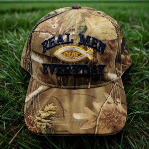 Op de afbeelding: Camouflage baseballpet met de tekst "REAL MEN PRAY EVERYDAY" in marineblauwe letters. Het woord "PRAY" staat boven een geel en wit vissymbool. De pet heeft een gebogen klep en een knoop aan de bovenkant.