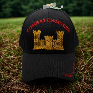 May include: Black baseball cap with "COMBAT ENGINEER" embroidered in red and a gold castle emblem. The cap has a curved brim and a tag attached to the top. The cap is sitting on a bed of grass.