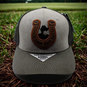 May include: A grey and black trucker hat with a brown leather patch showing a cowboy silhouette within a horseshoe. The hat has the text "CHILL BILLY" on the patch and "KAMEL ORIGINAL" on the brim.