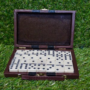 Puede incluir: Un juego de dominó marrón en un estuche abierto, sobre hierba verde. Los dominós son blancos con puntos negros. El estuche rectangular tiene un asa y un cierre dorado. El estuche mide unos 20 cm de largo.