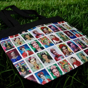 May include: Black tote bag featuring a repeating pattern of portraits of Frida Kahlo. The colorful images are set against a white background, with a black handle and trim. The bag is resting on a bed of green grass.