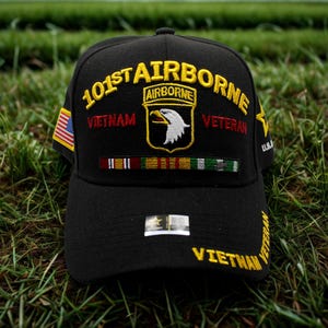 May include: Black baseball cap with embroidered text and patches. The cap features "101st AIRBORNE" in yellow, "VIETNAM" in red, and a patch with an eagle. An American flag patch is on the side. The cap is on a grassy surface.