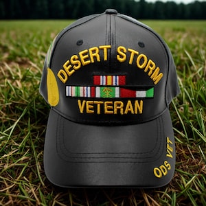 Puede incluir: Gorra de béisbol negra con la inscripción dorada "DESERT STORM VETERAN". La gorra presenta un parche con cintas militares y el texto "ODS VET" en la visera. La gorra está sobre hierba verde.