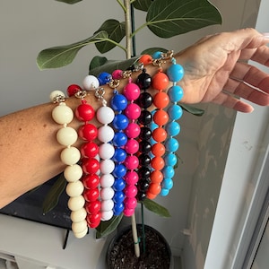 Puede incluir: Una colección de ocho pulseras de cuentas con esferas grandes y lisas en color crema, rojo, blanco, azul, rosa intenso, negro, naranja y azul claro. Cada pulsera está terminada con un cierre de palanca de metal en tono dorado.