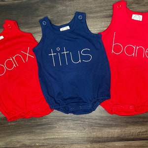 Puede incluir: Tres bodies de bebé sin mangas en rojo, azul y blanco con los nombres "banx", "titus" y "bane" bordados en blanco.
