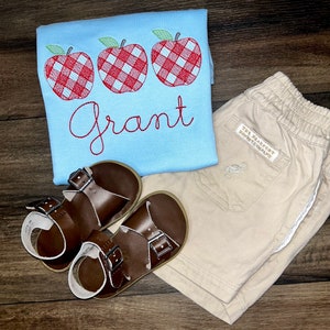 May include: A light blue t-shirt with three red and white gingham apples embroidered on the front. The name "Grant" is embroidered below the apples. A pair of brown leather sandals and beige shorts with a white stripe down the side are also pictured.