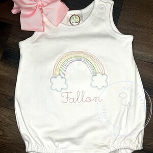 Pode incluir: Macacão branco para bebê com um arco-íris e nuvens bordados na frente. O nome "Fallon" está bordado abaixo do arco-íris. O macacão tem um laço rosa no ombro.