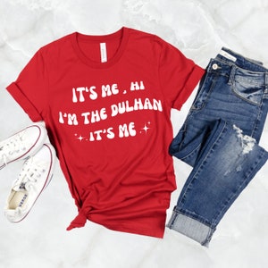 May include: Red t-shirt with white text that reads "It's me, hi I'm the dulhan It's me" with sparkling stars on either side of the last "It's me".