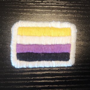 May include: A rectangular patch with a white border, featuring horizontal stripes of yellow, white, purple, and black. The patch is likely an emblem or symbol, possibly for a cause or identity.