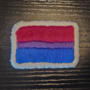 Op de afbeelding: Een rechthoekige patch met een witte rand en een ontwerp dat de biseksuele pride-vlag vertegenwoordigt. De vlag heeft drie horizontale strepen: een bovenste streep van rood, een middelste streep van paars en een onderste streep van blauw.