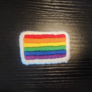 Puede incluir: Parche bordado con el diseño de la bandera del arcoíris. El parche tiene forma rectangular con un borde blanco y esponjoso. Los colores de la bandera incluyen rojo, naranja, amarillo, verde, azul y morado. Adecuado para ropa o accesorios.
