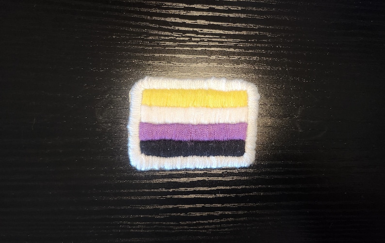 May include: A rectangular patch with a white border, featuring horizontal stripes of yellow, white, purple, and black. The patch is likely an emblem or symbol, possibly for a cause or identity.