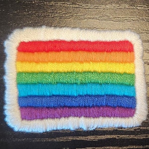 Pode incluir: Um patch retangular com um design de arco-íris, com listras horizontais em vermelho, laranja, amarelo, verde, azul e roxo. O arco-íris é delimitado por uma borda branca e fofa. Este patch é adequado para roupas ou acessórios.