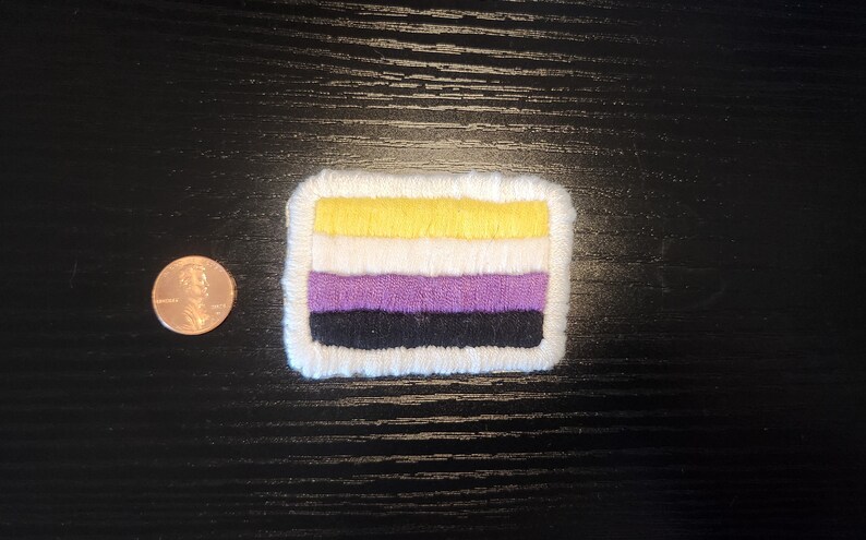 May include: A rectangular patch featuring the non-binary pride flag, with horizontal stripes of yellow, white, purple, and black, bordered by white stitching. A penny is next to the patch for size comparison.