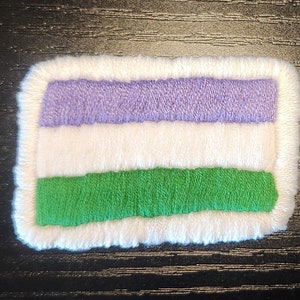 May include: Embroidered patch with a genderqueer pride flag design. The rectangular patch features horizontal stripes in lavender, white, and green, with a white border. The patch is made of fabric and has a textured appearance.