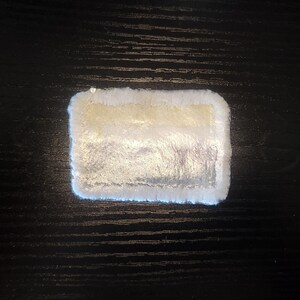 May include: A rectangular, plush coaster with a white, fluffy border and a shimmering, pearlescent center. The center has a gradient of colors, including silver and gold. The coaster is designed to protect surfaces from drink condensation.