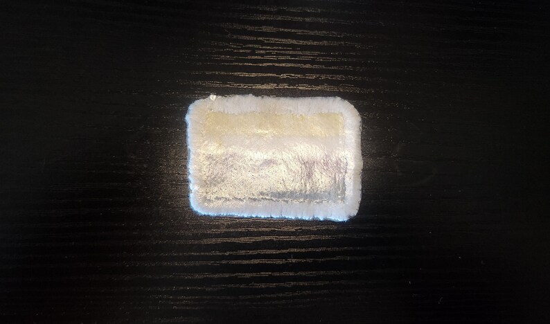 May include: A rectangular, plush coaster with a white, fluffy border and a shimmering, pearlescent center. The center has a gradient of colors, including silver and gold. The coaster is designed to protect surfaces from drink condensation.