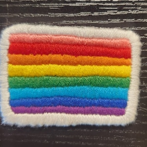 Puede incluir: Un parche rectangular con un diseño de arcoíris, con rayas horizontales en rojo, naranja, amarillo, verde, azul y morado. El parche tiene un borde blanco y esponjoso. Es probable que el parche sea un artículo bordado o cosido.