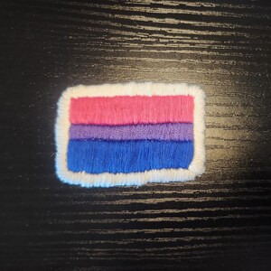 Pode incluir: Patch bordado com a bandeira do orgulho bissexual. O design apresenta listras horizontais em rosa, roxo e azul, com uma borda branca. Ideal para roupas ou acessórios.
