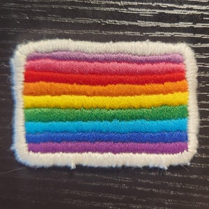 Pode incluir: Um patch retangular com um design de arco-íris, com listras horizontais em vermelho, laranja, amarelo, verde, azul e roxo. O patch tem uma borda branca e fofa. As cores são vibrantes e a textura parece macia.