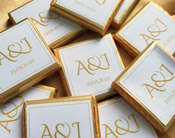 May include: A pile of gold-wrapped chocolate squares with white labels. Each label features the initials "A&I" and the date "25.05.2025" in gold lettering. The chocolates are arranged on a gold-colored surface.