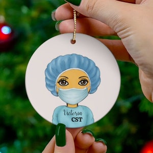May include: A white ceramic Christmas ornament featuring a cartoon illustration of a person wearing a blue surgical cap, a blue surgical gown, and a blue face mask. The text "Victoria CST" is printed below the illustration.