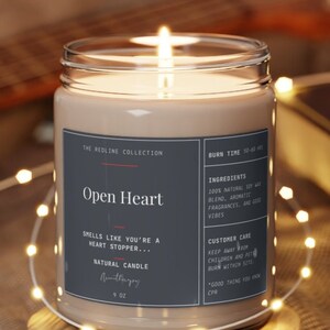 Peut inclure: Une bougie en cire de soja naturelle dans un bocal en verre transparent avec une étiquette noire qui indique "The Redline Collection Open Heart" et "Smells like you're a heart stopper... Natural Candle". La bougie est posée sur un dessous de verre en bois.