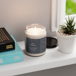 May include: A gray candle with the text "The Surgeon" and "Smells like the same thing every time in 30 minutes or less. Natural Candle" on the label. The candle is lit and the flame is visible.