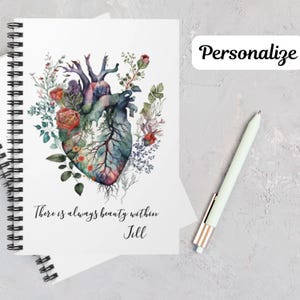 May include: A white notebook with a spiral binding features a watercolor illustration of a human heart with flowers and vines growing from it. The text "There is always beauty within Jill" is printed below the illustration.