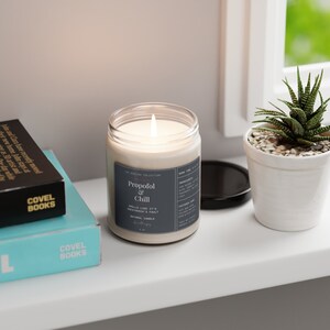 May include: A white candle with a black label that reads "The Pirlim Collection Propofol & Chill" and "Smells like it's anaesthesia's fault. Natural Candle." The candle is lit and sitting on a white surface.