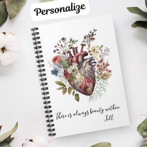 May include: A white spiral bound notebook with a watercolor illustration of a human heart surrounded by flowers. The text "There is always beauty within Jill" is printed below the illustration.