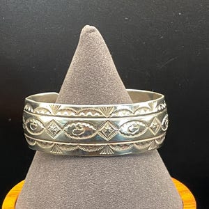 May include: A silver cuff bracelet featuring a wide band with intricate stamped patterns, including repeating diamond shapes, sunburst motifs, and scalloped edges along the top and bottom borders of the polished metal surface.