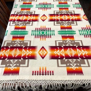 May include: A cream-colored woven blanket featuring a repeating geometric pattern in shades of red, orange, yellow, green, and brown. The design includes diamond and cross motifs with a fringed edge along the bottom and side borders.