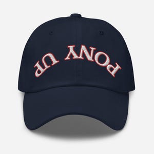 May include: A navy blue baseball cap with white embroidered text that reads "PONY UP" in a curved arc. The text is outlined in red.