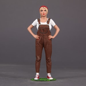 May include: A hand-painted figurine of a woman wearing a white shirt, brown overalls, a red bandana, and red shoes. The figurine is standing on a green base.
