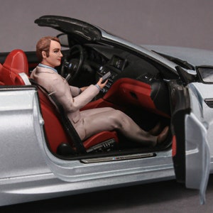 May include: A silver convertible car with the door open and a person sitting in the driver's seat. The car has red interior accents and a black top.