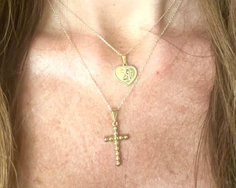 May include: Two gold necklaces with pendants. The top necklace has a heart-shaped pendant with an angel design. The bottom necklace has a cross pendant with small beads.