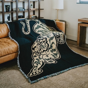 May include: A black and gold woven throw blanket with a tiger design. The blanket is draped over a brown leather couch in a living room setting.