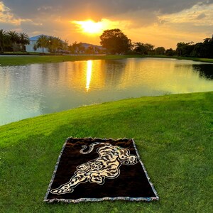 May include: A brown and black woven throw blanket with a tiger design. The blanket is laid out on a grassy area near a lake with a sunset in the background.